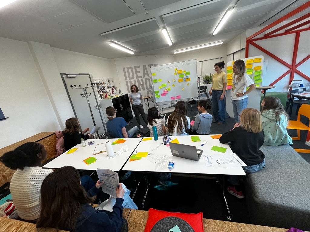 Students in a workshop review ideas on sticky notes and a whiteboard in a bright classroom.